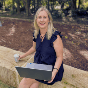 Anne-Marie sitting on a sandstone block in a park, drinking coffee while working on a laptop